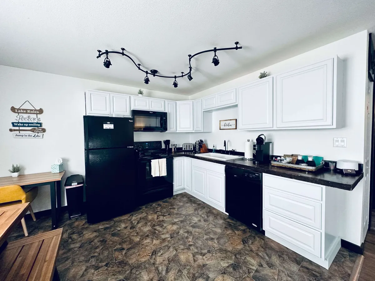 A bright kitchen scene featuring crisp white cabinetry, black appliances, and thoughtfully arranged countertop decor. The space is styled to feel both functional and inviting, hinting at possibilities like hosting dinner parties, baking, or enjoying a morning coffee together.