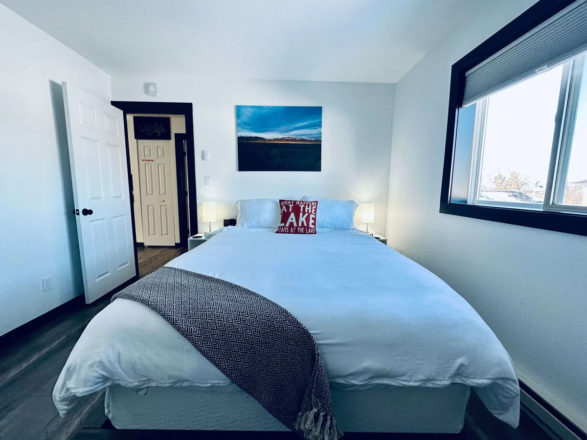 The image presents another bedroom with crisp white linens, lake-themed artwork above the bed, glowing nightstand lamps, and a cozy decorative throw blanket. The mood is calm and inviting, evoking the sense of a peaceful retreat after a day’s adventure. Ample natural light and subtle touches create a soothing ambiance.