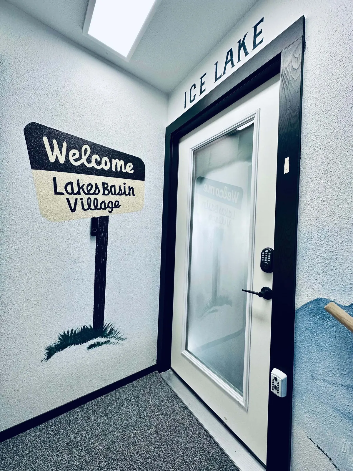 The image displays a friendly 'Welcome' sign situated next to the door of Ice Lake Condo, providing a clear sense of entry. Soft natural lighting filters across the doorway, inviting visitors into the space and setting a warm tone for arrival.