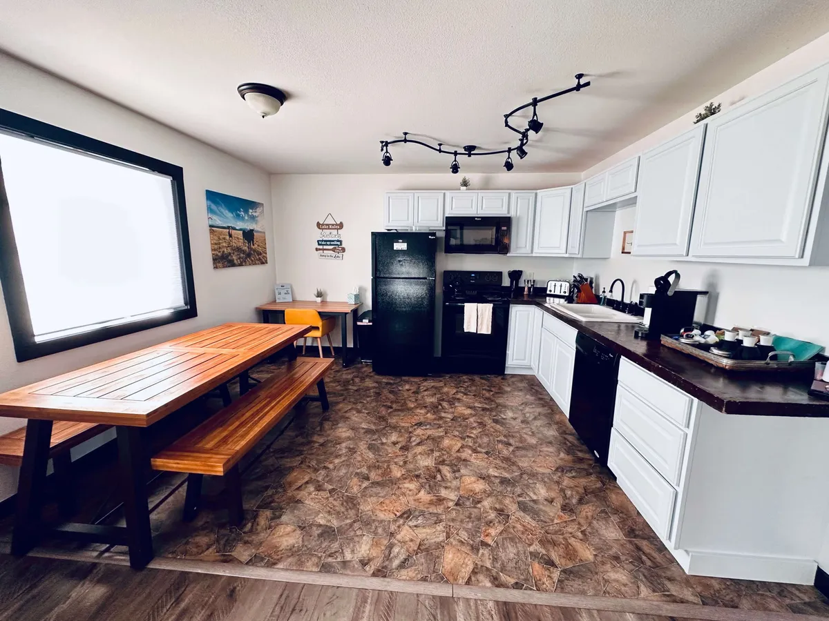 This cozy scene shows a dining table set beside a black refrigerator and adjacent kitchen counter, framed by a window that brings in natural light. The layout emphasizes a warm, inviting atmosphere, ideal for family dinners, small gatherings, or relaxed cooking sessions.