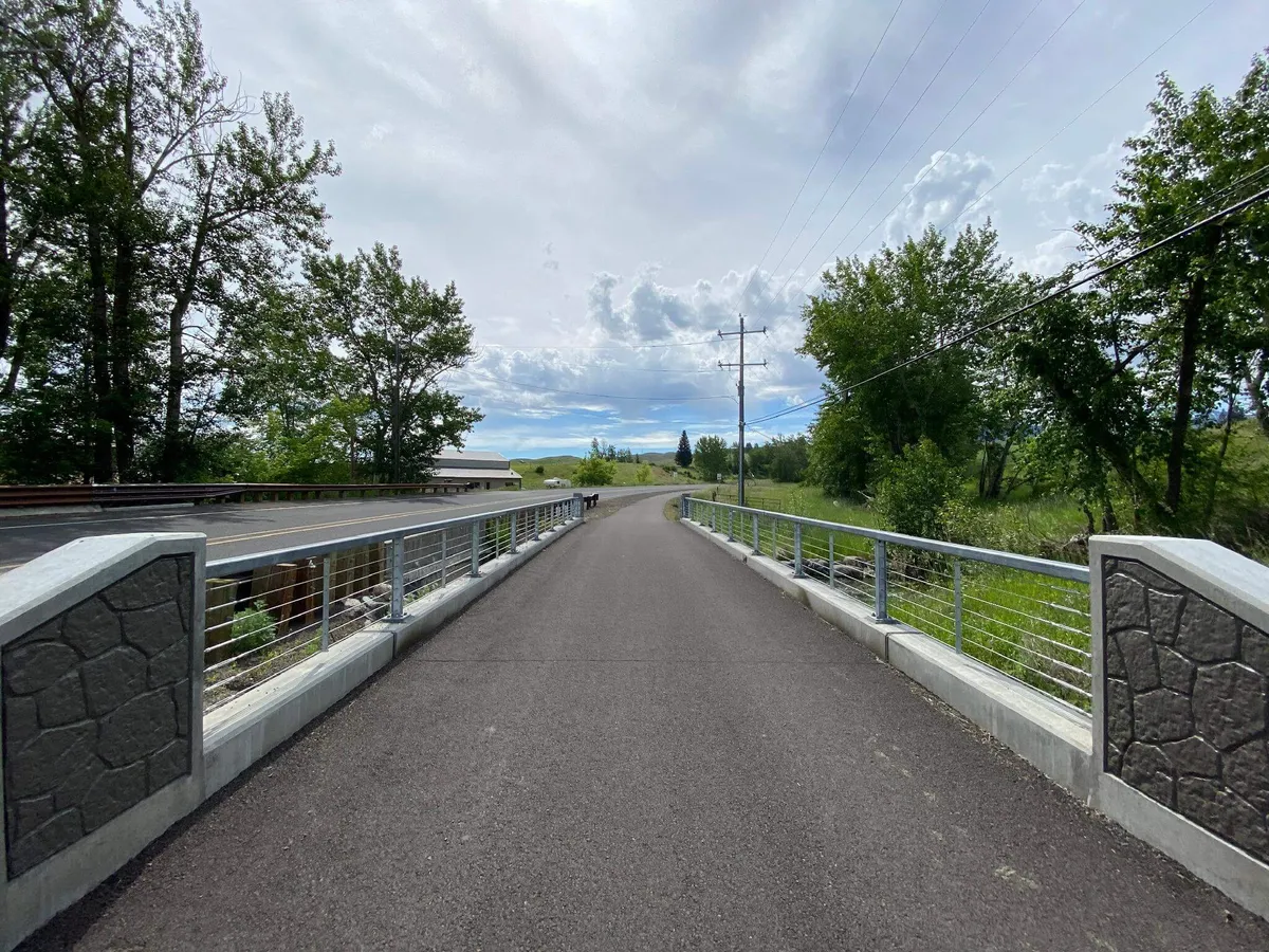 A picturesque exterior view of a bridge and pathway winding through lush trees under an open sky, inviting guests to walk, bike, or explore the surrounding natural landscape. The tranquil scene hints at the outdoor adventures that await right outside the property.