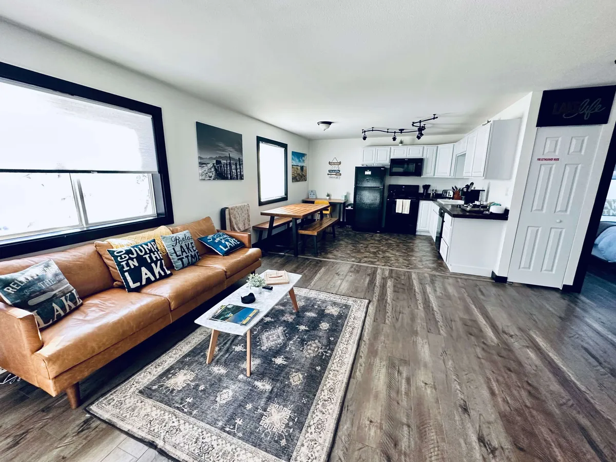 A sunlit living area featuring a plush leather sofa adorned with decorative pillows, a coffee table, and a dining table with views into the kitchen area. Large windows fill the space with natural light and offer glimpses of the surrounding trees, creating a serene and cozy atmosphere. The layout invites gathering, relaxation, and social activities.