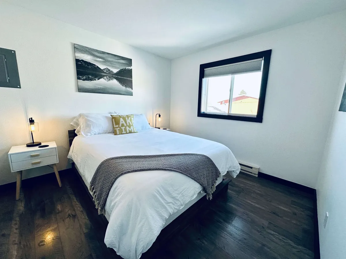 This image features a cozy bedroom bathed in soft natural light, with a plush bed outfitted in fresh linens, decorative pillows, tasteful artwork on the wall, and a window offering a glimpse of the outdoors. The décor is serene and inviting, creating an atmosphere perfect for relaxation and restful sleep.