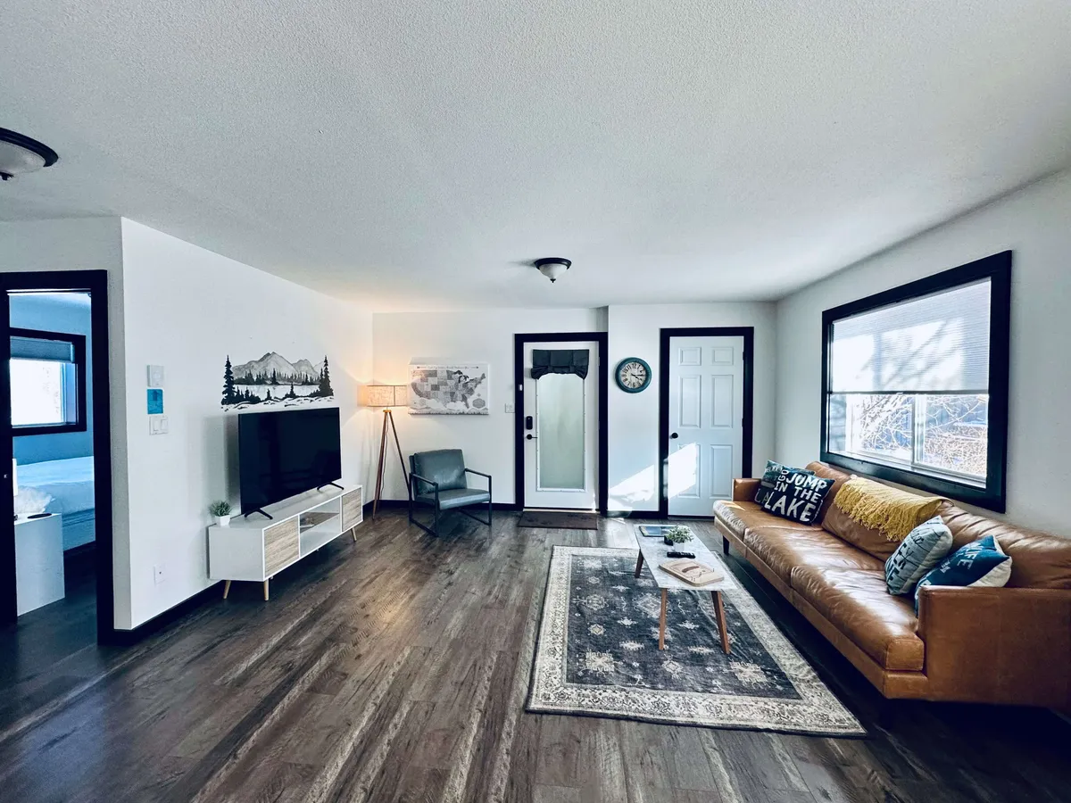 The image showcases a cozy living room featuring a comfortable sofa, a stylish rug, a flat-screen television, a warm lamp, and inviting artwork on the walls. Large windows let in natural light, creating a welcoming space perfect for relaxing, socializing, or unwinding with a cup of coffee.